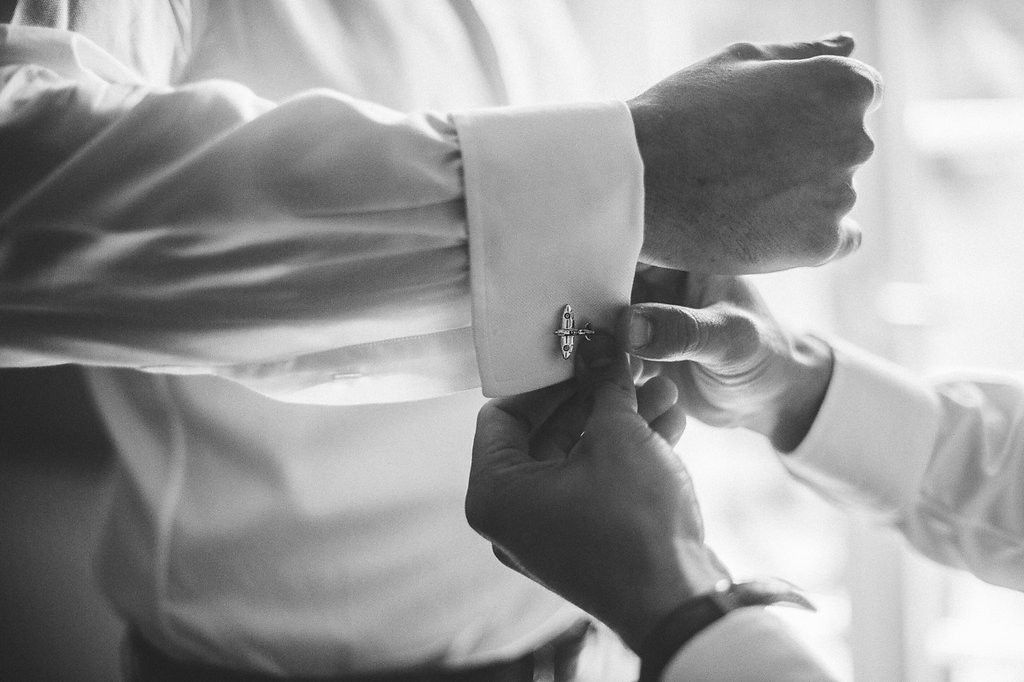 Black and white photo of cufflinks being fastened on groom's white dress shirt.