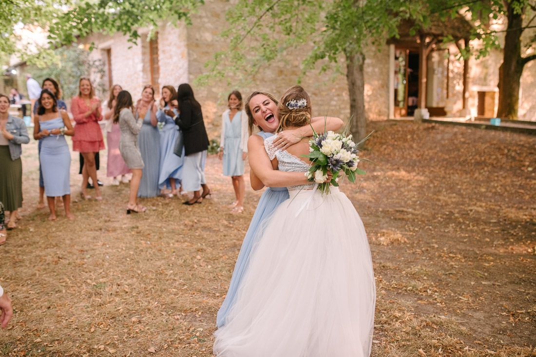 Bride in white dress sharing joyful hug with guest holding bouquet at sunny outdoor wedding celebration.