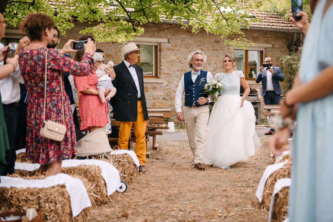 Bride walks down outdoor aisle flanked by hay bales and guests at rustic countryside wedding ceremony.