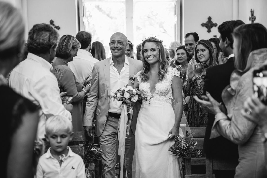 Bride walks down church aisle with escort in black and white wedding ceremony photo.