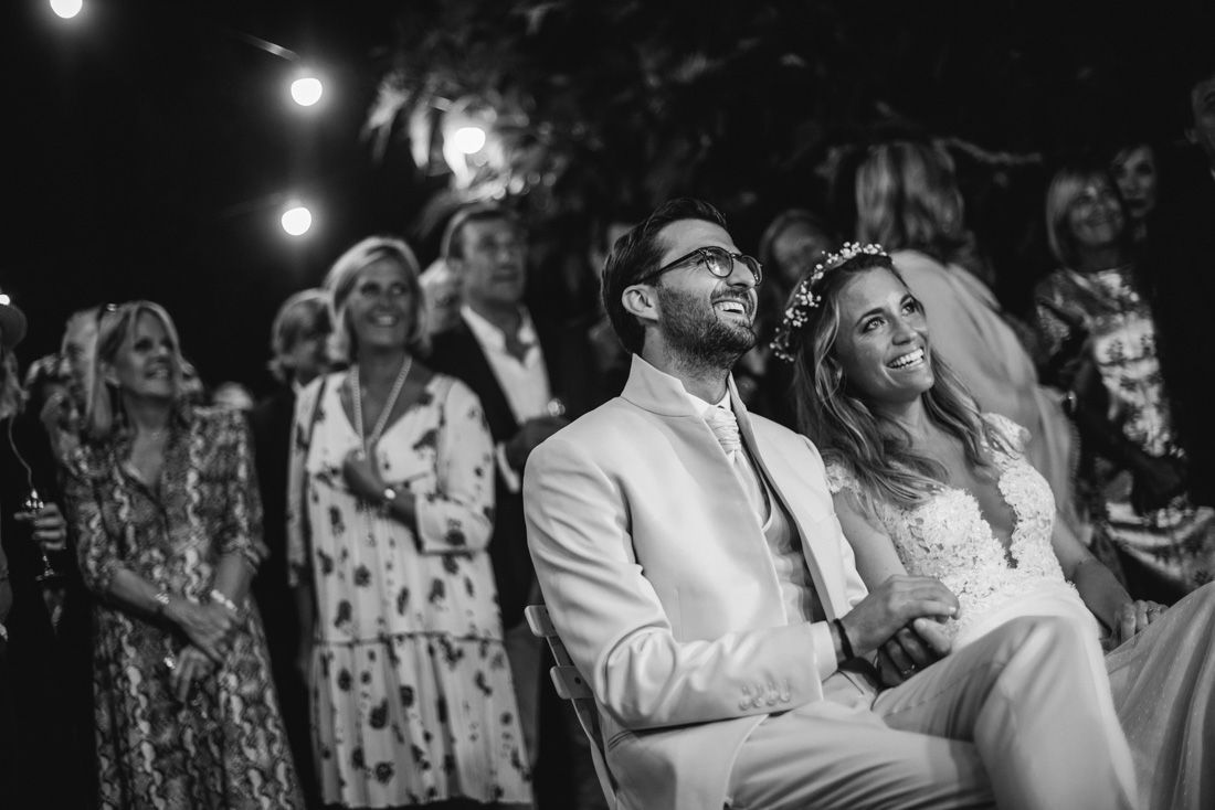 Black and white photo of a laughing bride and groom seated at their outdoor wedding ceremony.