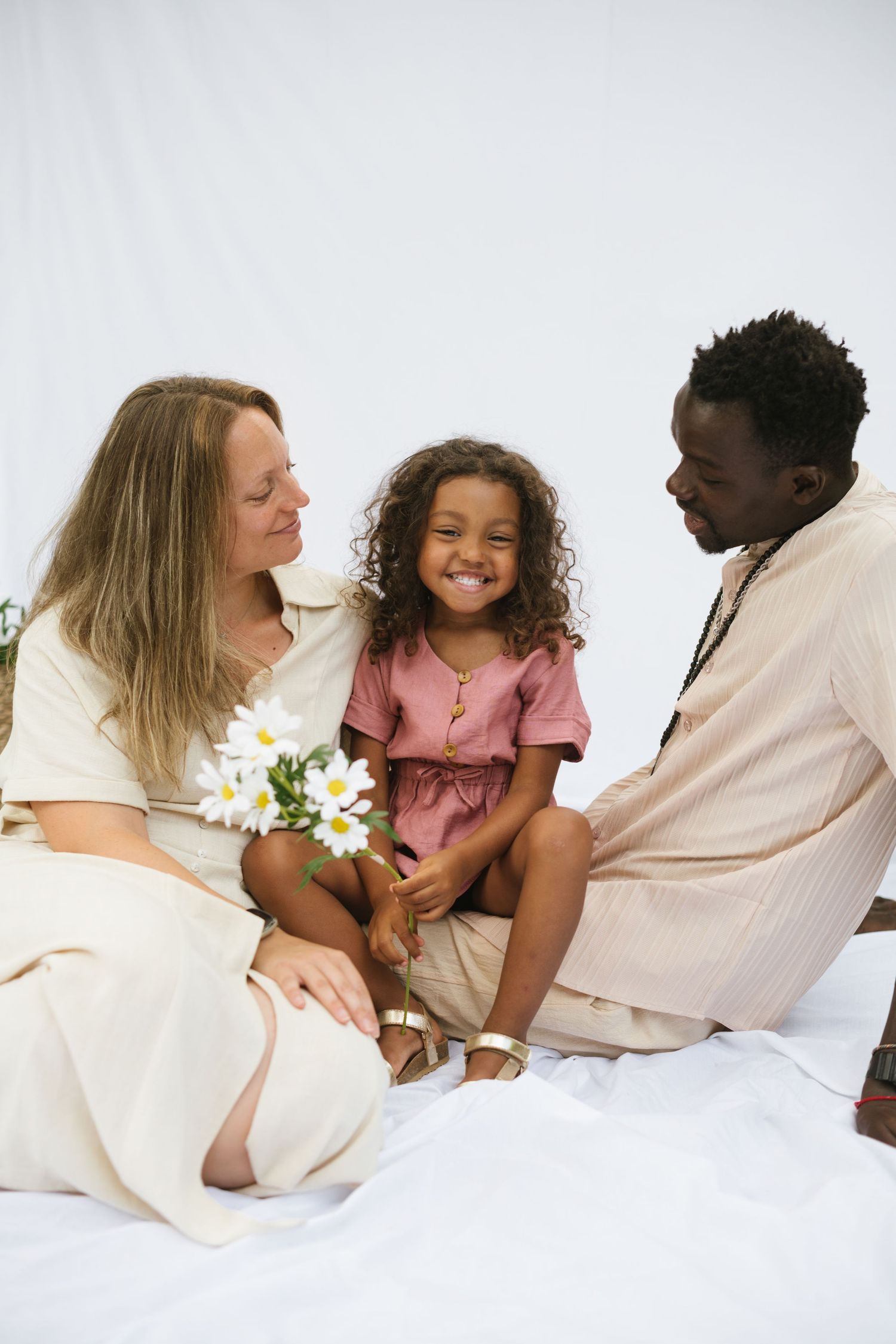 An interracial family sits together on white fabric as their curly-haired daughter holds white daisies.