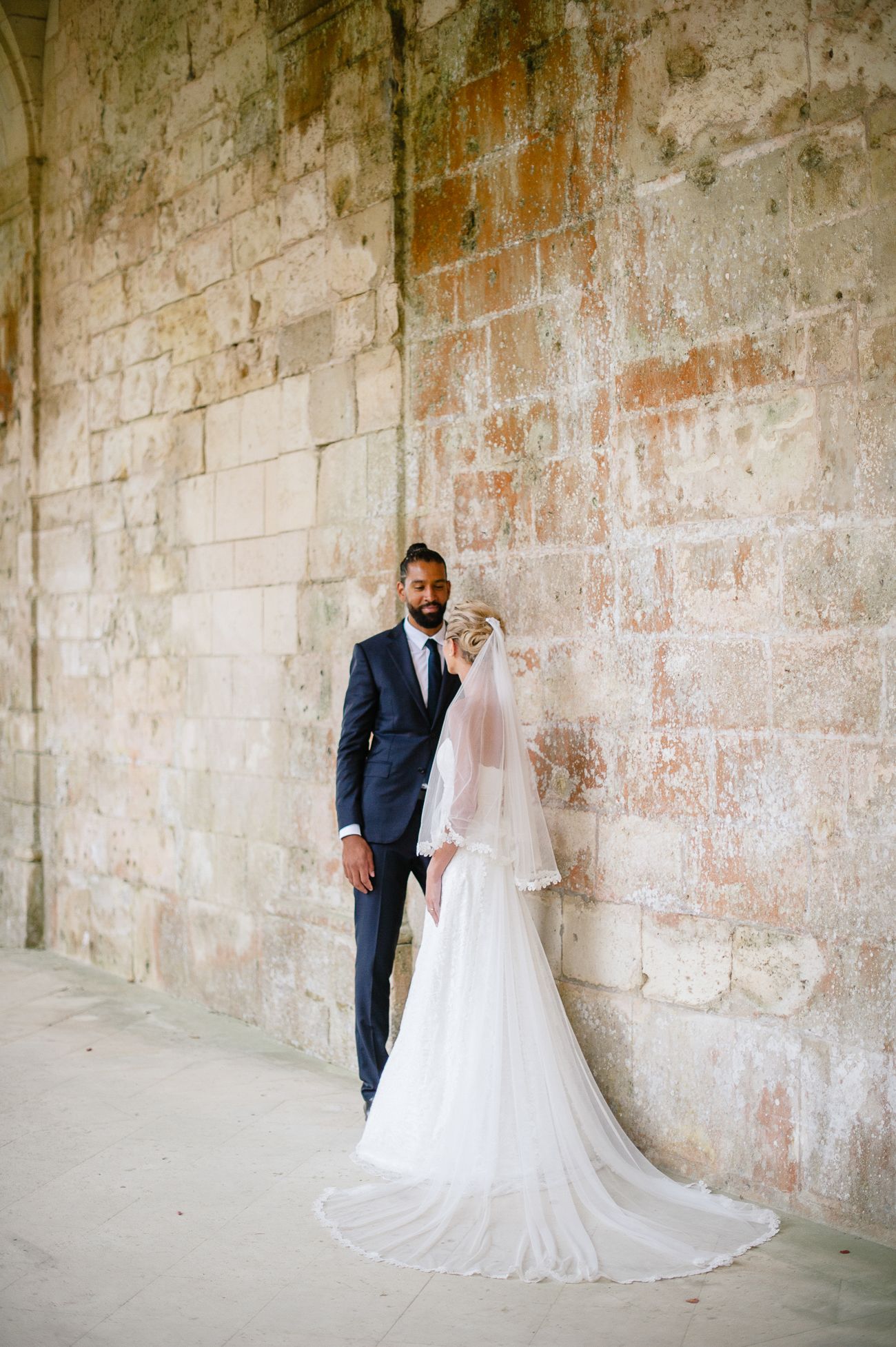 Bride and groom portrait against rustic stone wall, bride in flowing white gown with long veil, groom in navy suit.