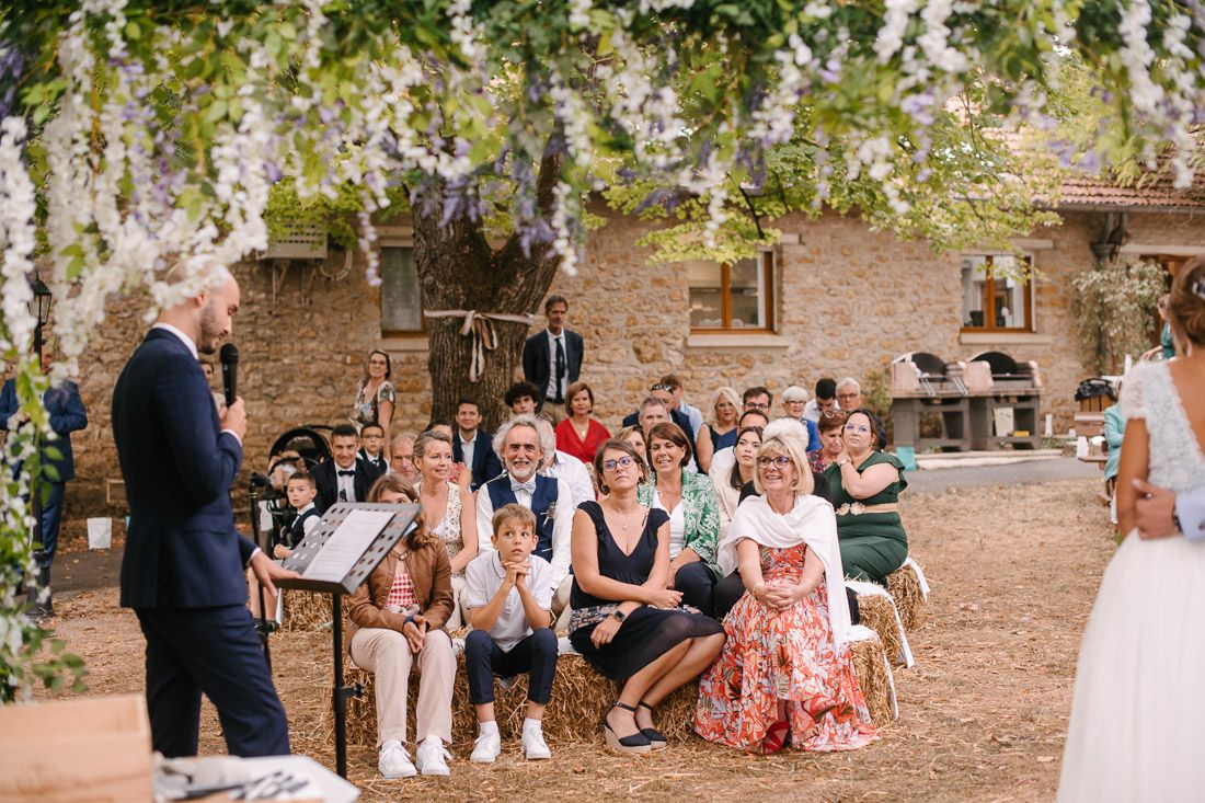 Outdoor wedding ceremony with guests seated under wisteria-draped pergola at rustic stone venue.