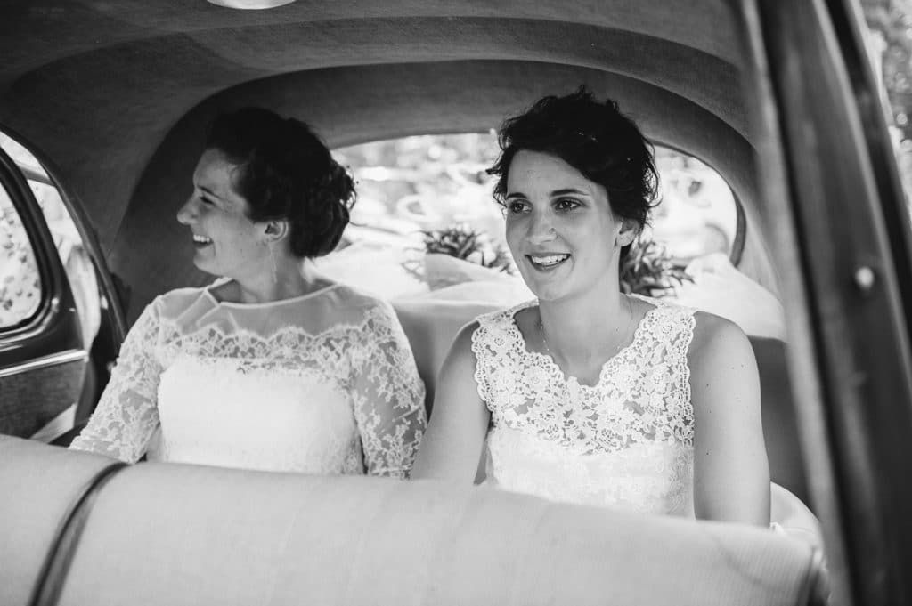 Two brides in lace wedding gowns smile together in the backseat of a vintage car.
