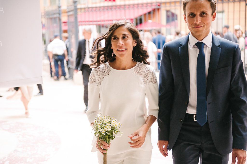 Bride holding white bouquet walks with groom in navy suit on sunny city street.