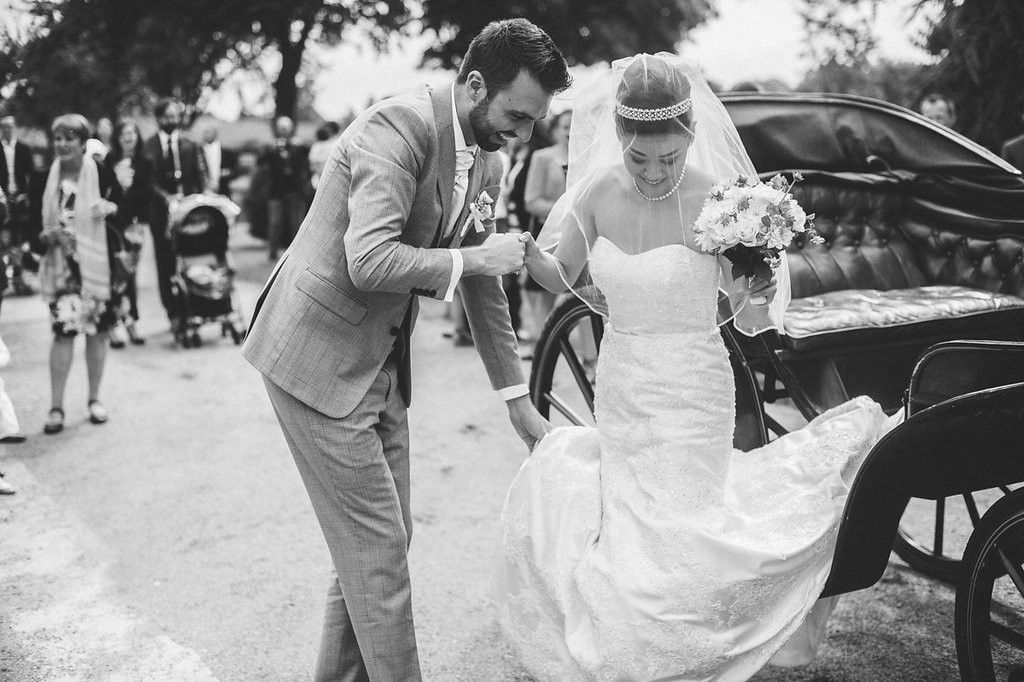 Groom in grey suit helps bride in white lace gown exit vintage black carriage at outdoor wedding.