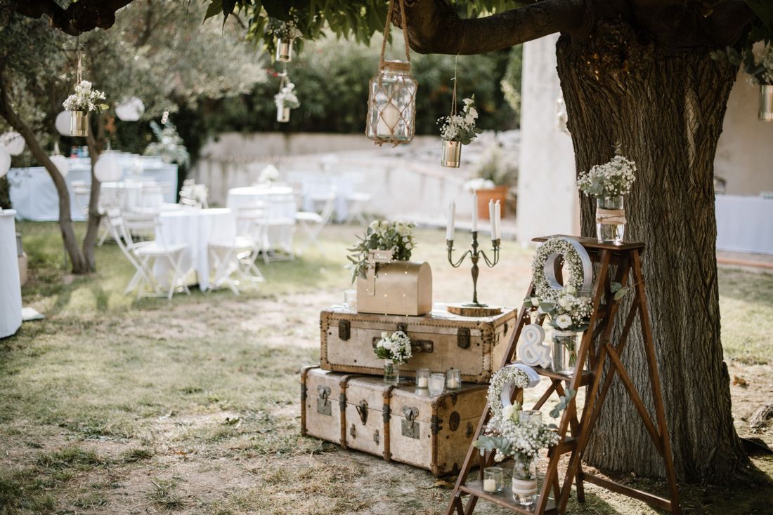 Rustic outdoor wedding decor with vintage suitcases, wooden ladder, candelabra, and floral arrangements under a tree.