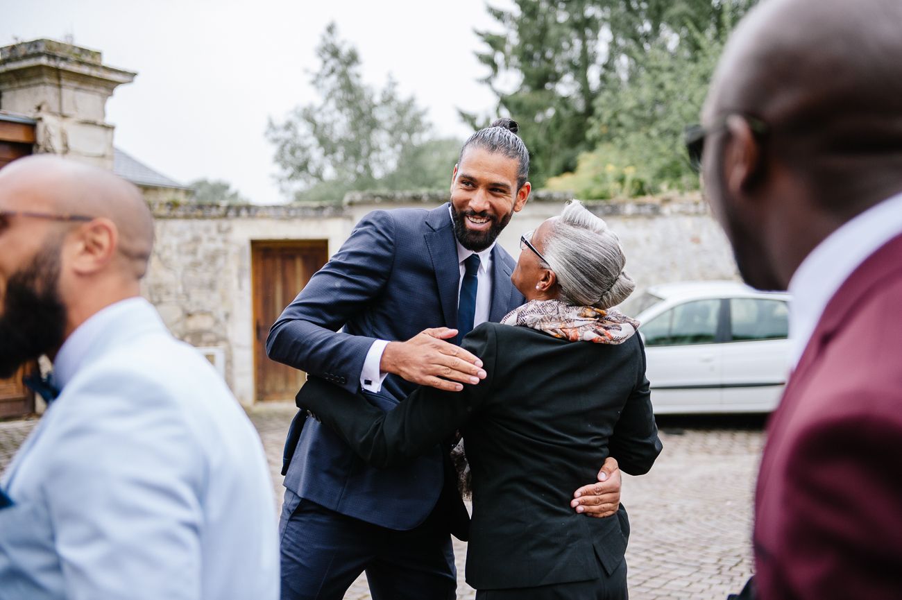 Two men in suits warmly embracing at an outdoor event, with guests in the background near a stone wall.