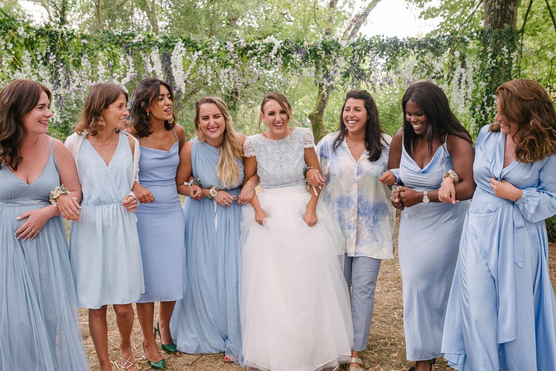 Bride surrounded by seven bridesmaids in matching light blue dresses laughing outdoors at wedding.