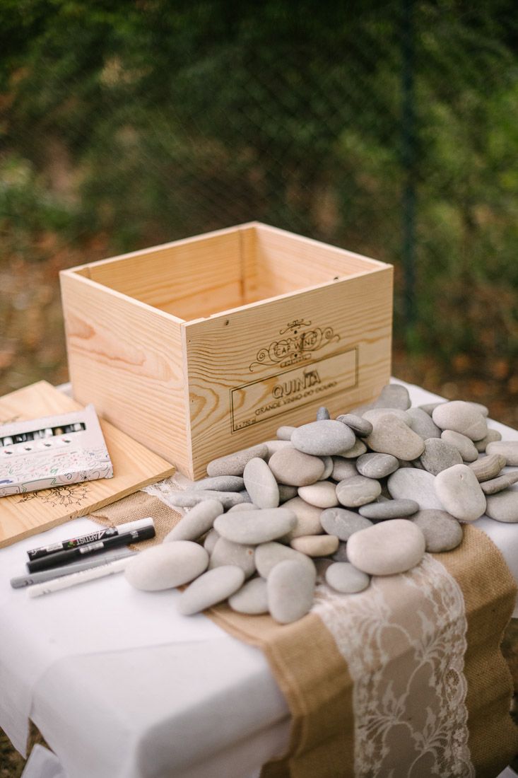 Rustic wedding guest book table with smooth gray stones, markers, and a wooden crate on lace-covered table.