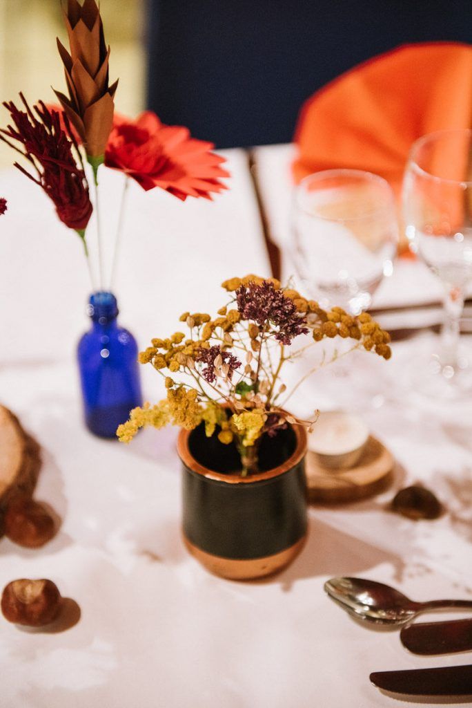 Wedding table decor with dried yellow flowers in a dark copper-rimmed vase and a blue bottle with red gerbera.