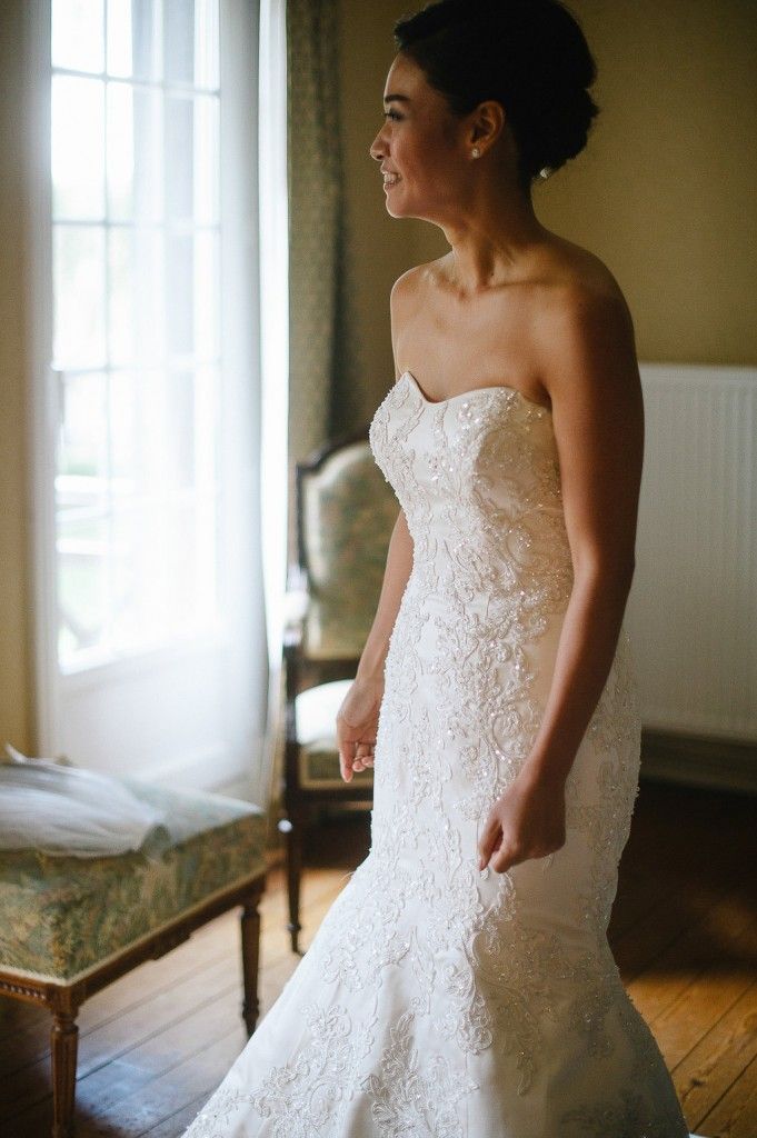 Elegant bride in strapless lace mermaid gown standing by bright window in vintage room.