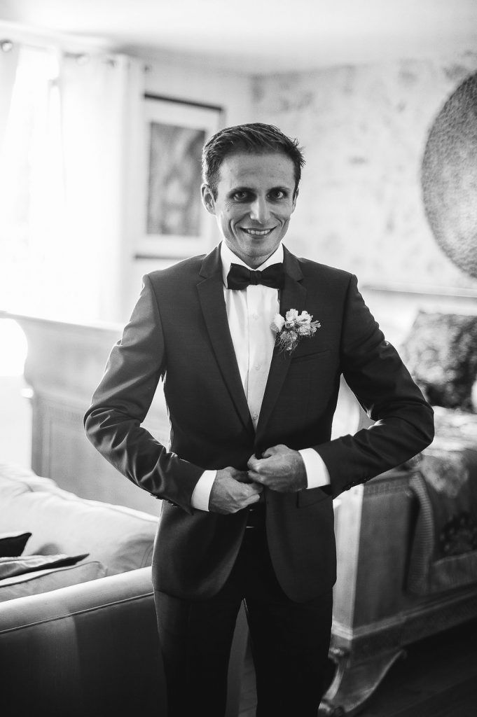 Smiling groom in black tuxedo and bow tie posing in elegant room before wedding.