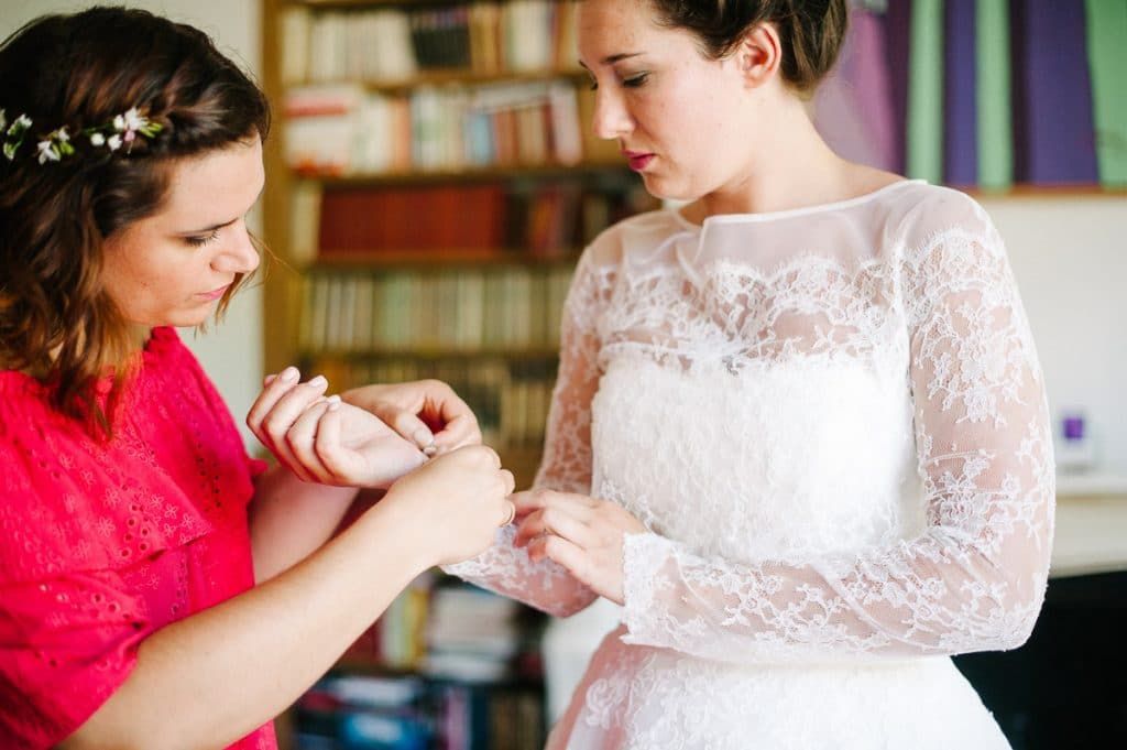 A bridesmaid in red helps a bride in a lace long-sleeve wedding dress fasten her cuff in a cozy room.