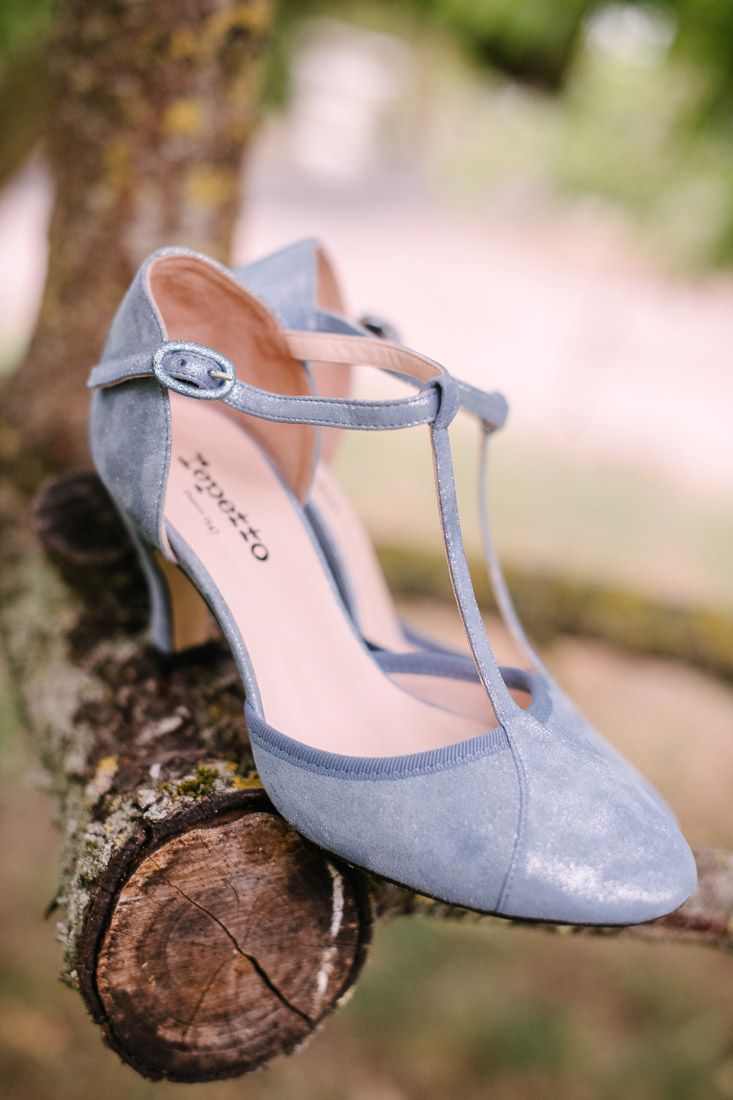 Elegant blue suede T-strap heels with ankle buckle displayed on a tree branch outdoors.