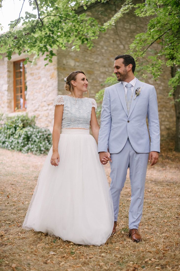 Bride in two-piece white gown and groom in light blue suit hold hands walking outside rustic stone venue.