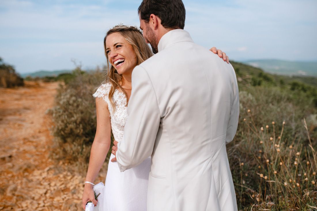 Joyful bride in white dress laughing with groom in beige suit on a sunny outdoor trail.