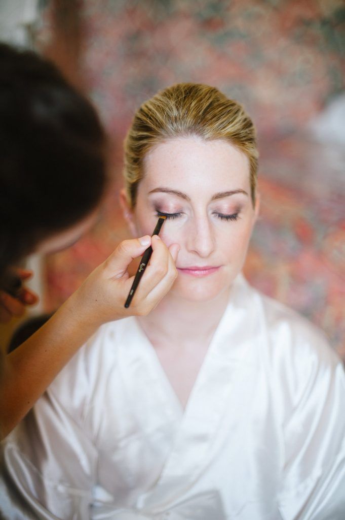 Professional makeup artist carefully applying eyeliner to a bride in a white robe.
