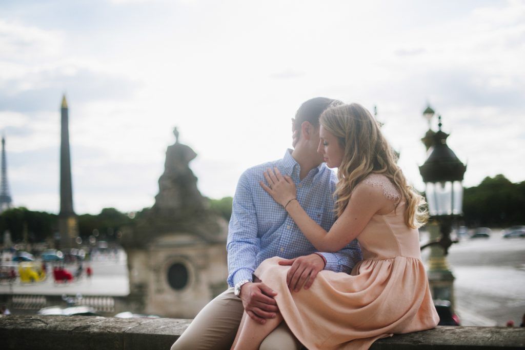Couple sharing a tender kiss while sitting on a stone ledge with the Eiffel Tower in Paris background.