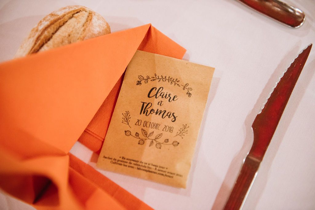Rustic kraft paper wedding place card for Claire and Thomas beside an orange napkin and bread roll.