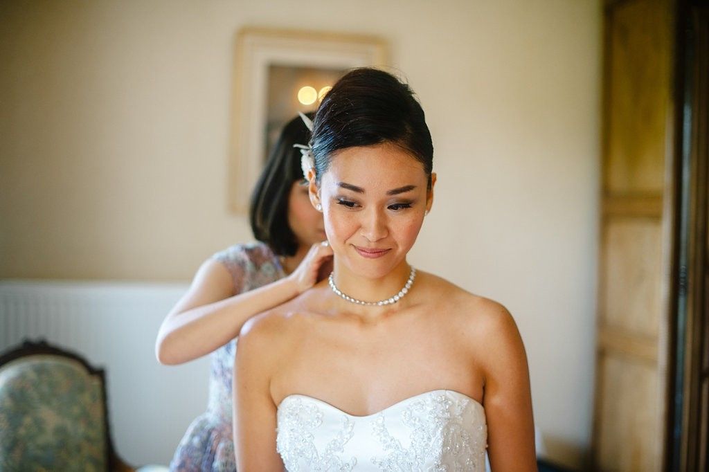 Bridesmaid adjusting earring on Asian bride in strapless lace gown with pearl necklace.