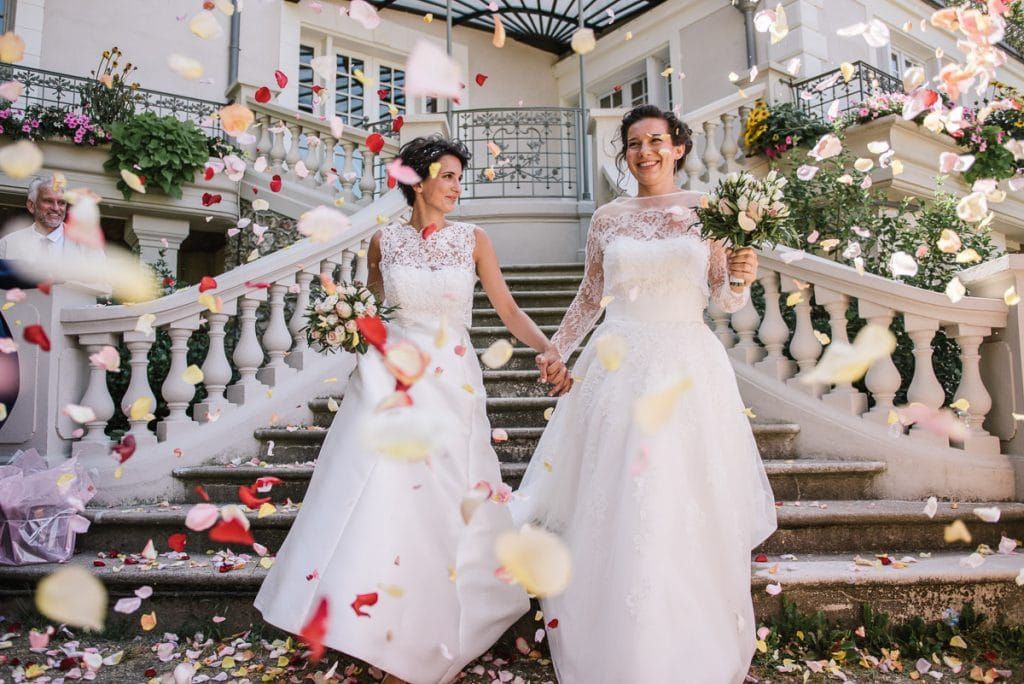 Two brides holding hands showered with colorful flower petals on grand staircase of French chateau wedding.
