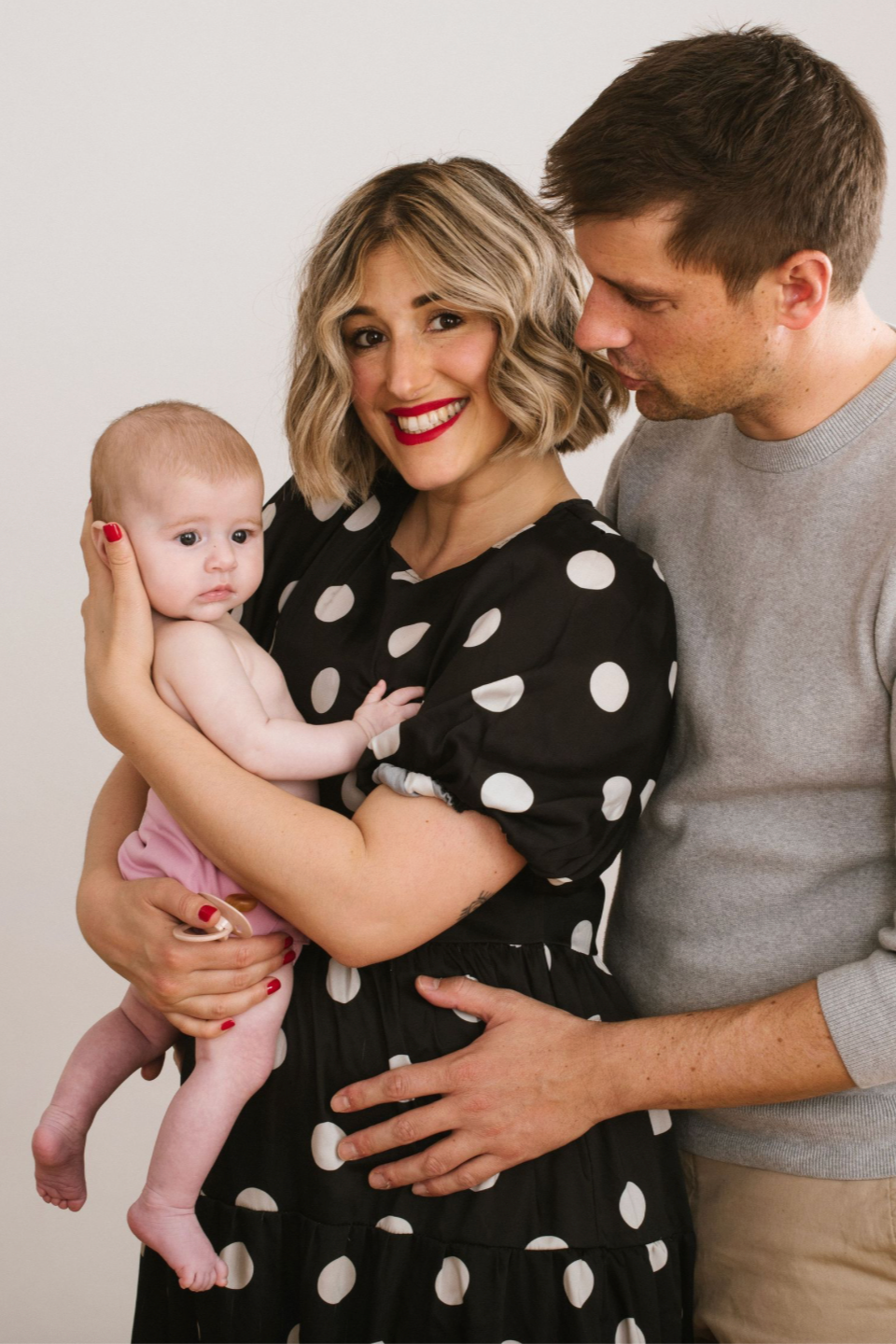A smiling mother in polka dot dress holds her newborn while father kisses her head in studio portrait.