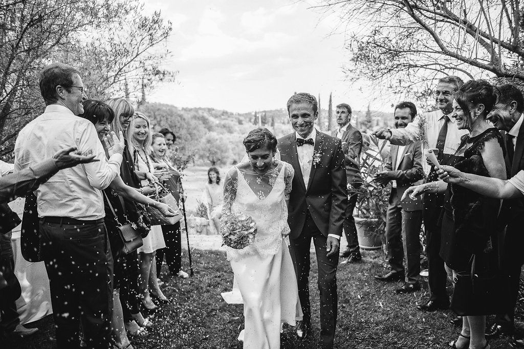 Black and white image of bride and groom walking through confetti-throwing guests at outdoor wedding celebration.