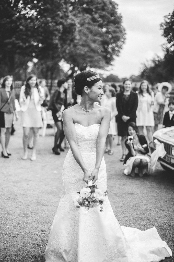 Beautiful bride in strapless lace gown and crystal tiara holds white bouquet while guests gather in background.