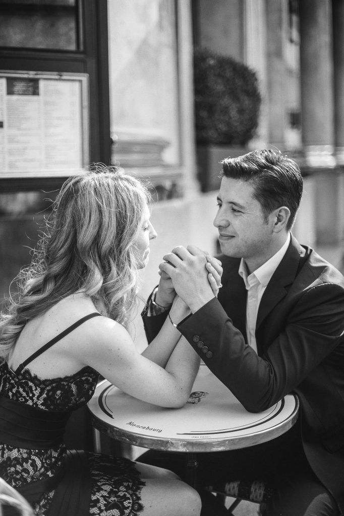 Black and white photo of couple holding hands across a small cafe table, gazing into each others eyes.