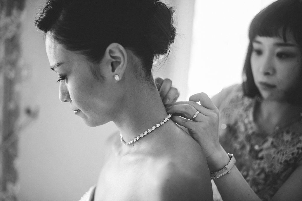 Woman helping bride fasten pearl necklace in black and white wedding preparation photo.