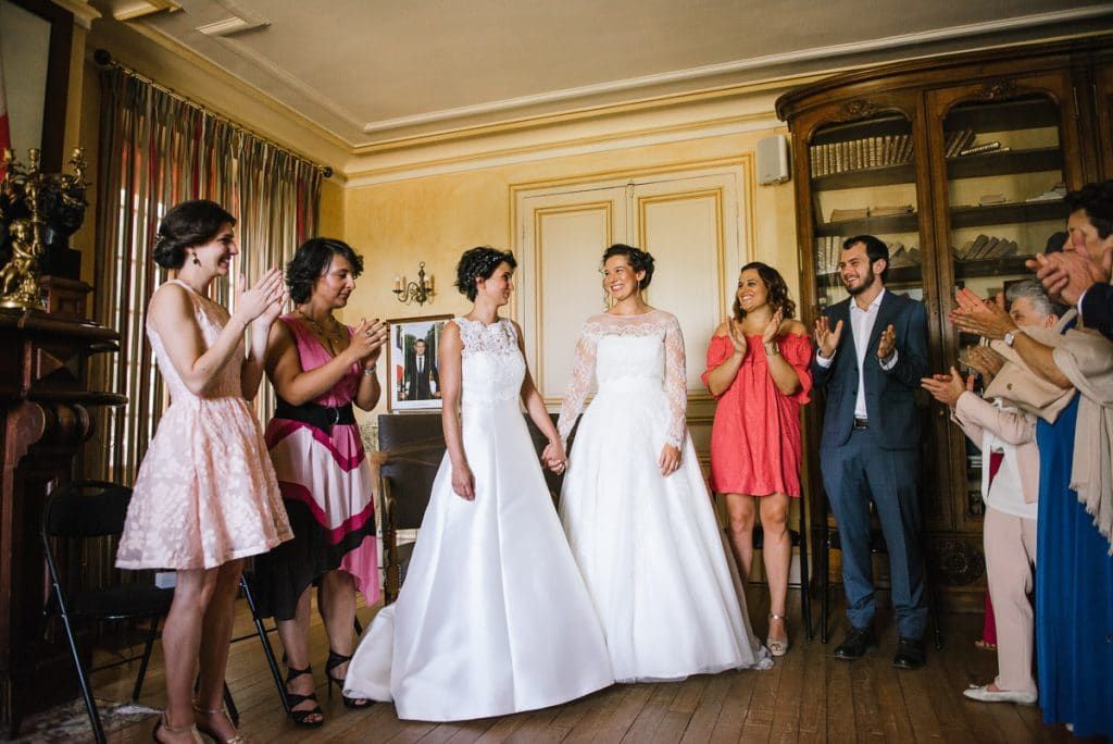 Two brides in white gowns celebrate with wedding guests clapping and cheering in elegant chateau interior.