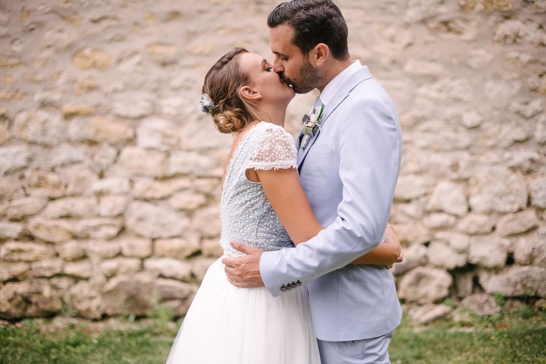 Bride and groom share romantic kiss in front of rustic stone wall after wedding ceremony.