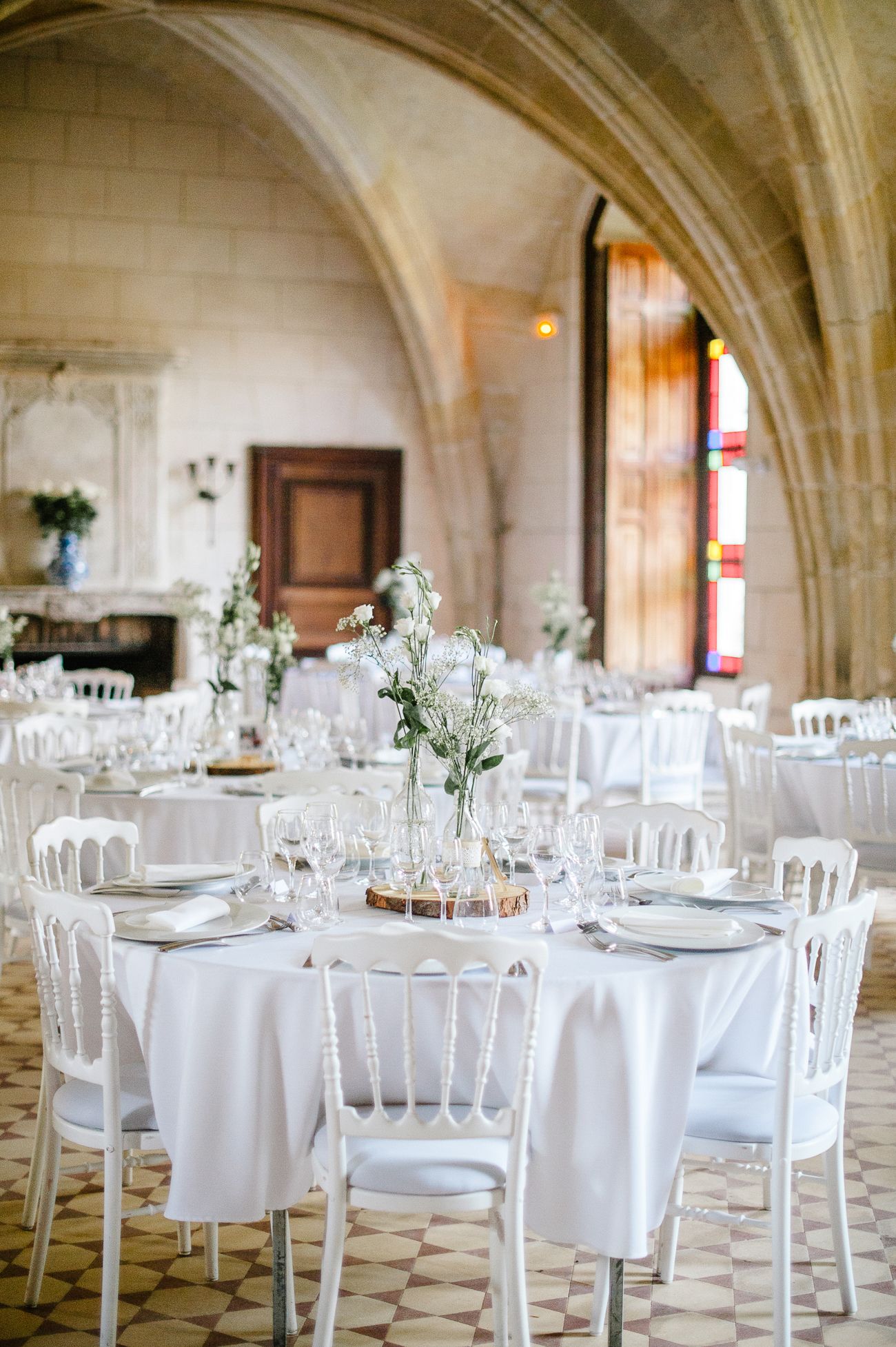 Elegant wedding reception hall with white round tables, chiavari chairs, floral centerpieces, and Gothic stone arched ceilings.