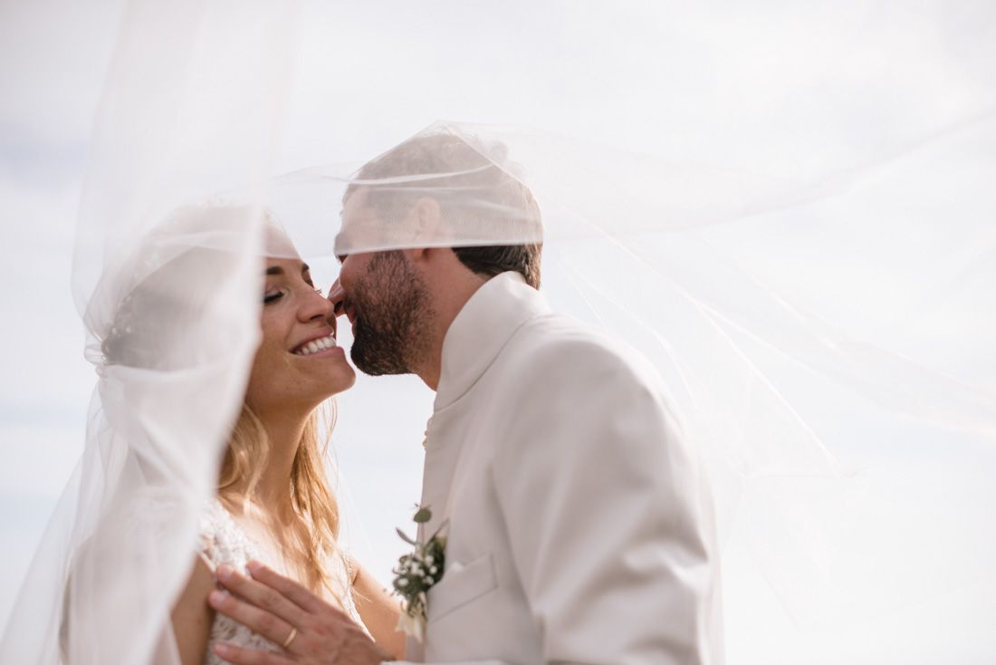 A bride and groom share a romantic kiss under a flowing white veil on their wedding day.
