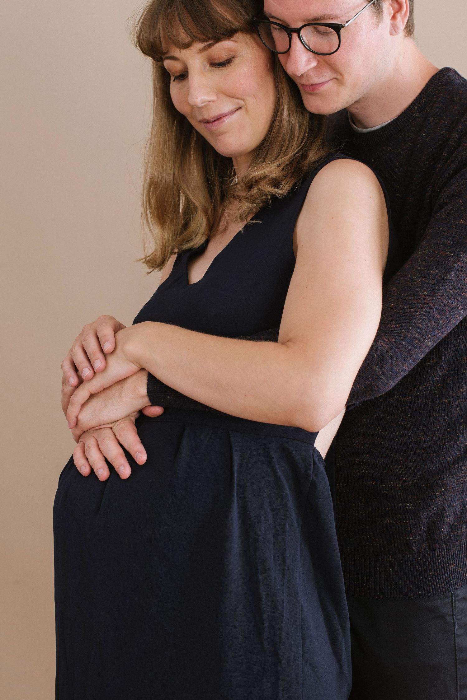 Couple sharing tender moment during maternity shoot, man embracing pregnant woman in black dress from behind.