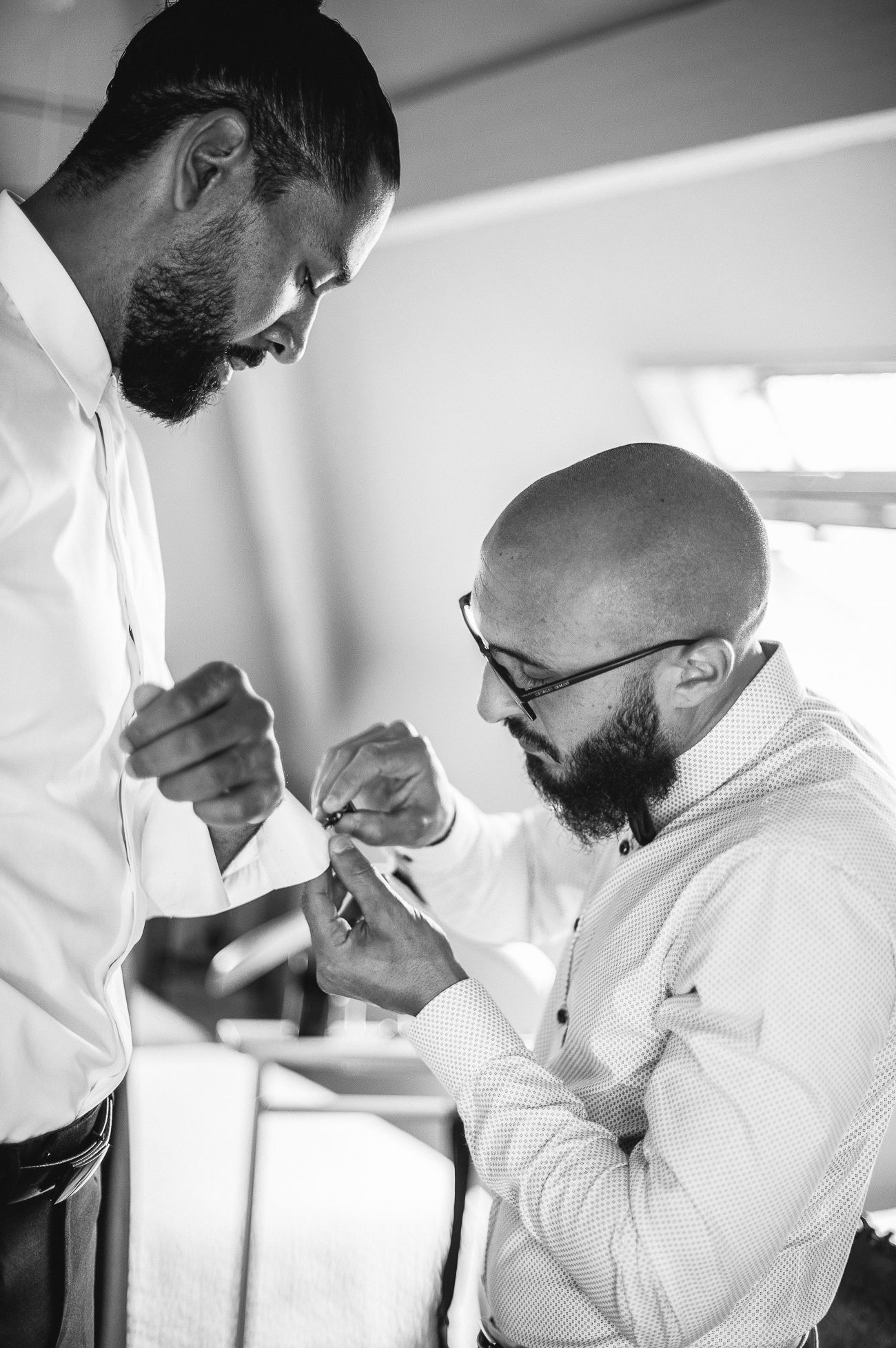 Black and white photo of two bearded men in dress shirts, one helping the other with cufflinks before a wedding.