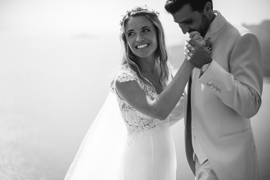 Black and white wedding photo of a groom kissing bride's hand outdoors, bride smiling in lace dress with veil.