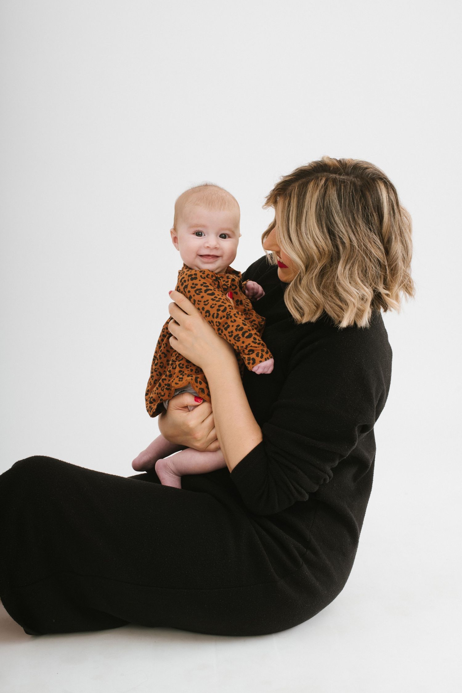 A blonde mother in black dress holds her smiling baby in a leopard print outfit on white background.