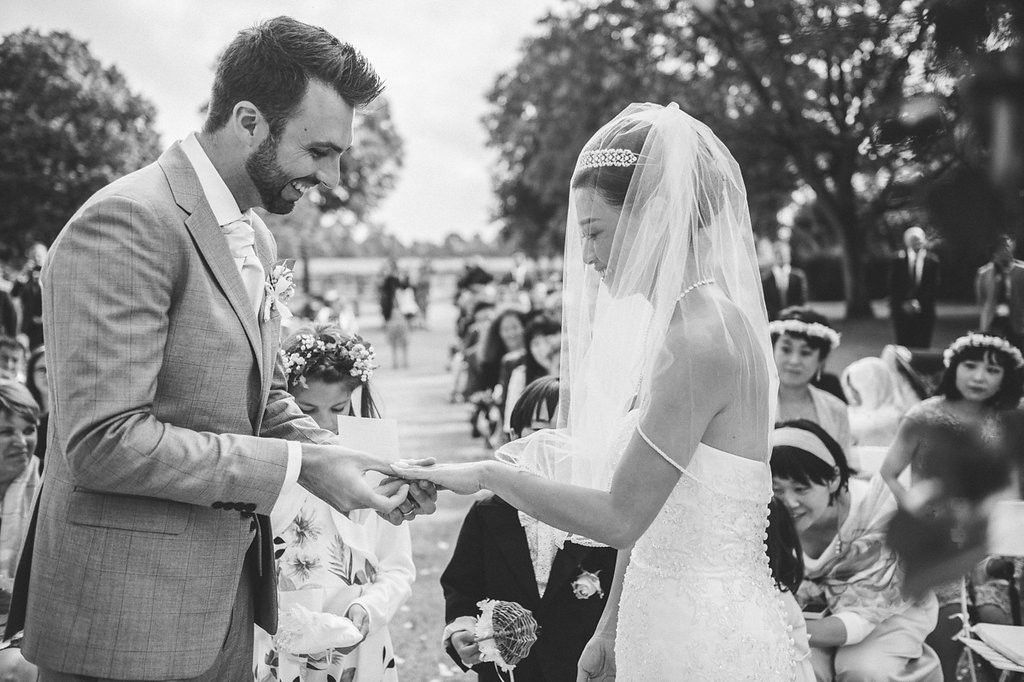 Bride places wedding ring on grooms finger during an intimate outdoor ceremony surrounded by seated wedding guests.