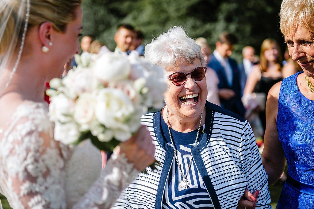 Elderly grandmother laughs joyfully as bride presents white rose bouquet at outdoor wedding.