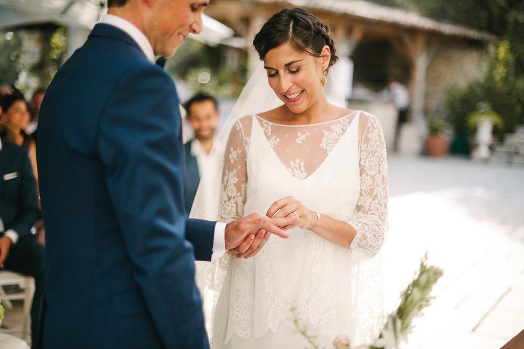 Bride smiling down as groom slides wedding ring onto her finger during outdoor ceremony.