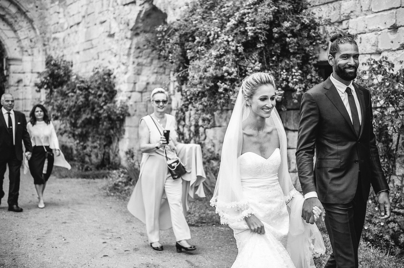 Black and white photo of bride and groom walking with wedding guests along a stone wall path.
