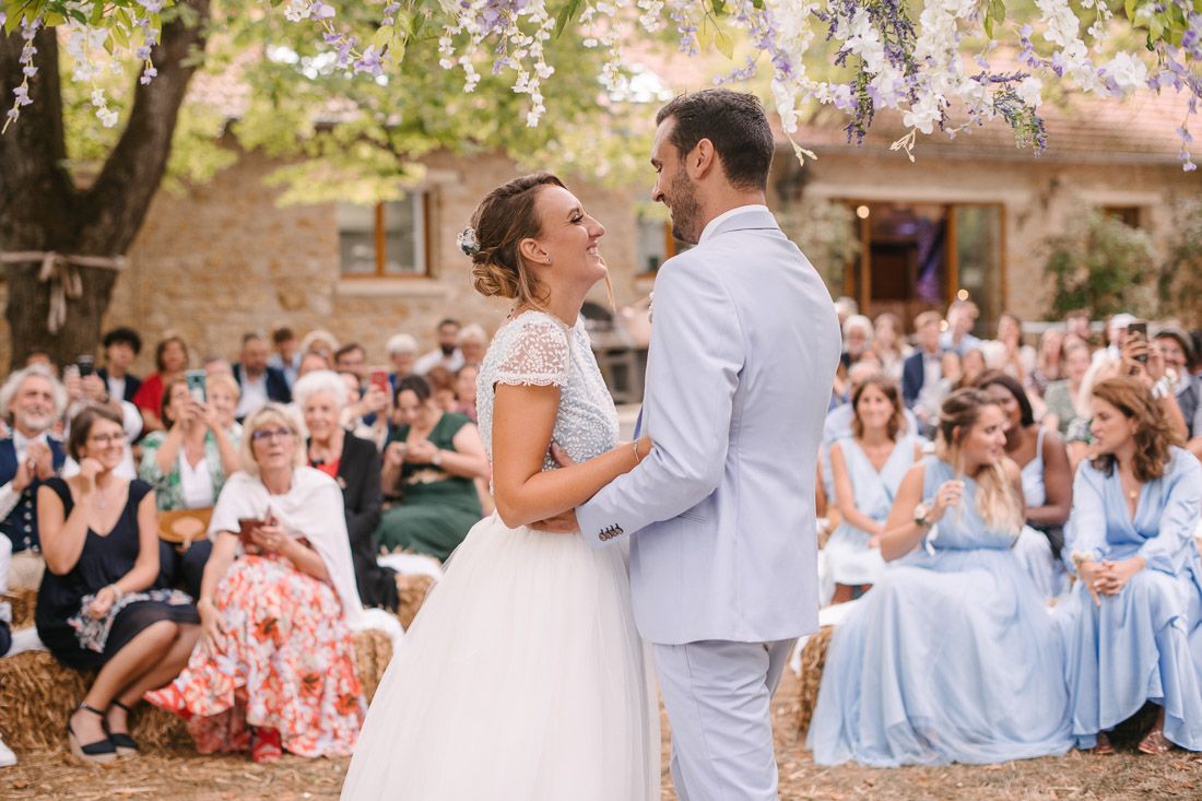 Bride and groom share first dance at outdoor wedding ceremony with guests seated around them under blooming trees.