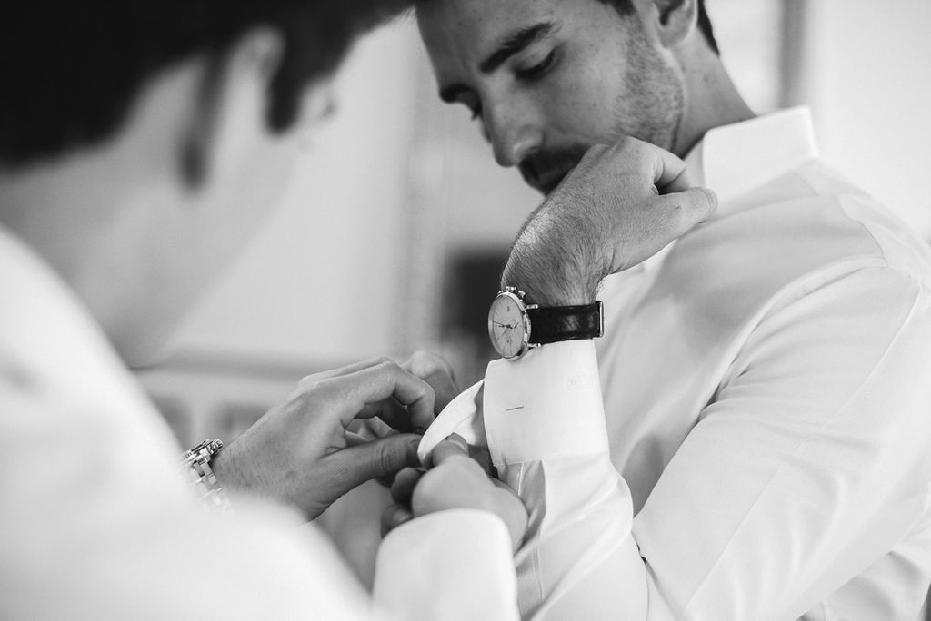 Groomsman helping groom adjust cufflinks in black and white wedding preparation photo.
