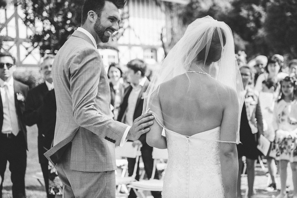 Smiling groom holds brides hands during outdoor wedding ceremony with guests seated in background.