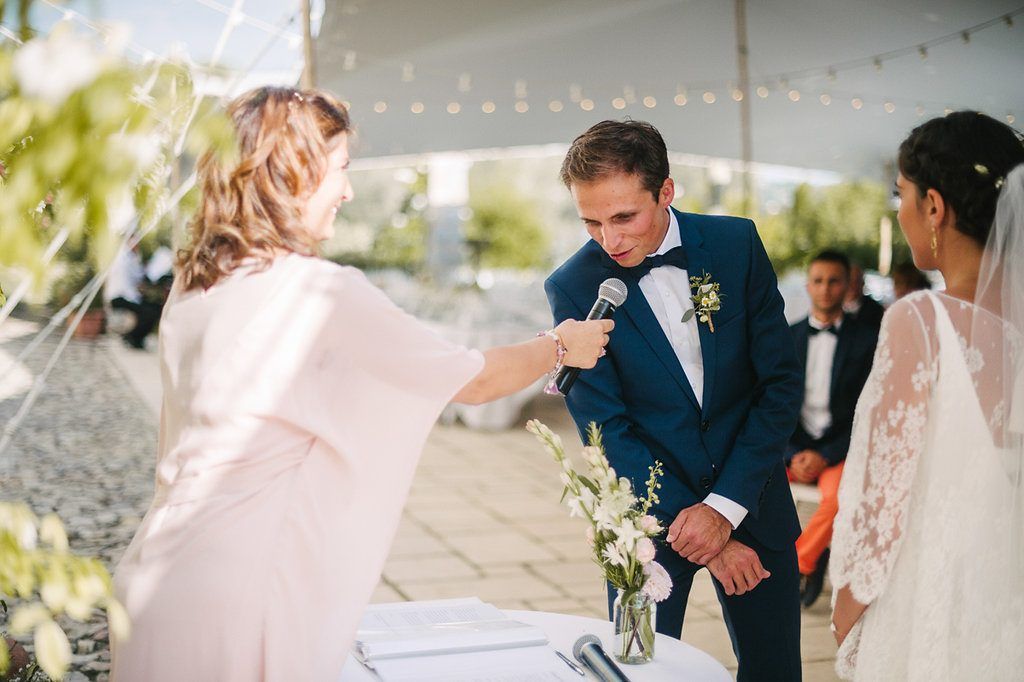 Officiant holds microphone for groom reading vows at elegant outdoor wedding ceremony under white tent.