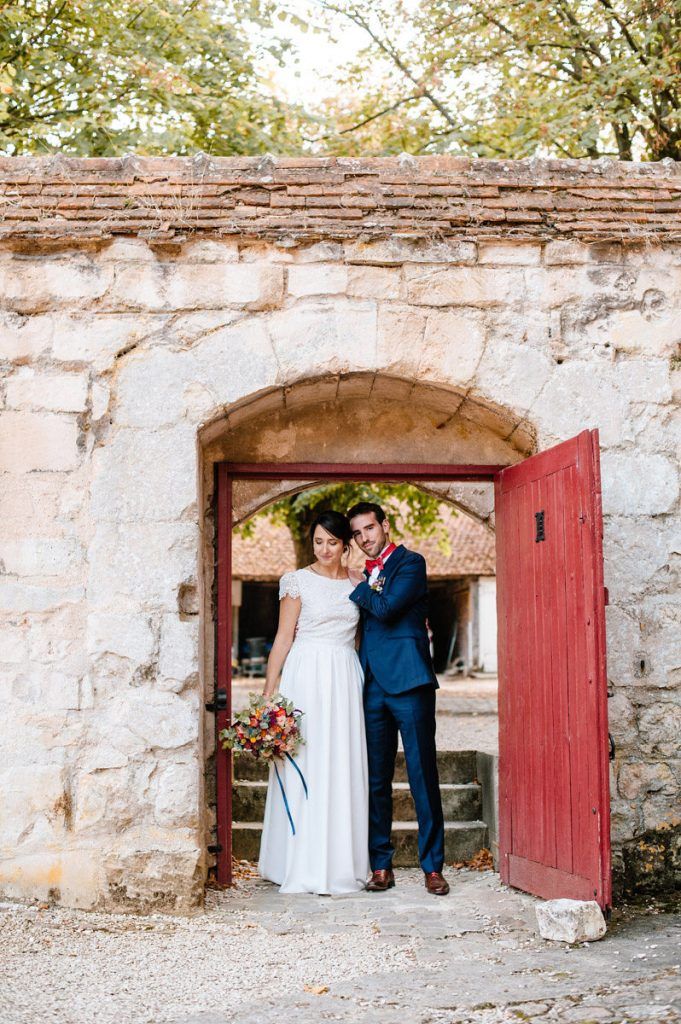 Bride and groom stand together in stone archway with red door holding colorful bouquet.
