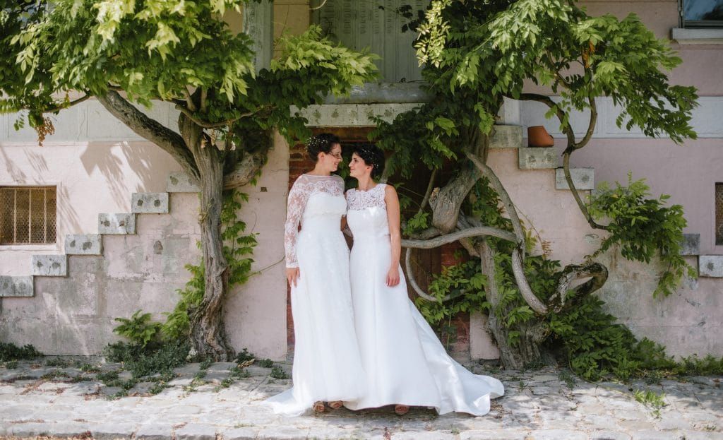 Two brides in elegant white wedding dresses posing together beneath a large tree outdoors.